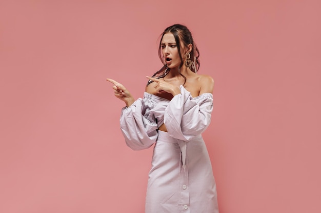 Disappointed slim woman with curly dark hairstyle in earrings trendy blouse and long lilac skirt pointing to place for text