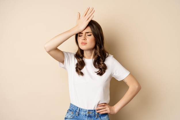 Disappointed woman reacting on failure, slap forehead, facepalm with sad face, beige background