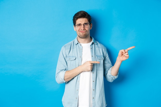 Disappointed young man in glasses pointing fingers right at copy space, showing promo banner and smirking dissatisfied, judging bad product, standing over blue background