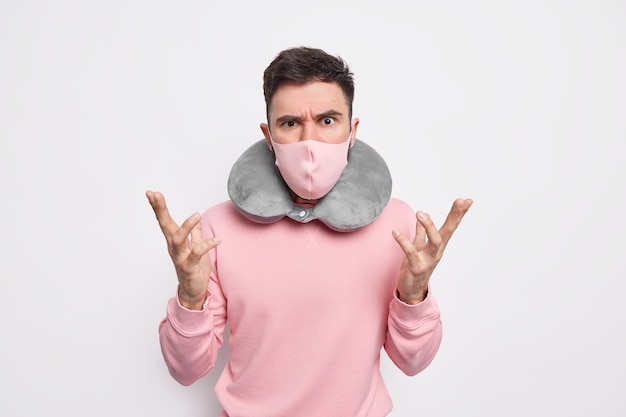 Discontent indignant man raises hands looks angrily and disappointed wears comfortable neck pillow for traveling in public transport protective mask to prevent coronavirus spread. Tourism concept