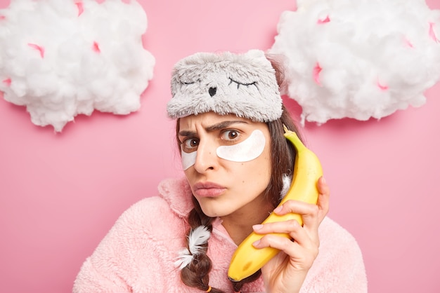 Discontent woman looks angrily at camera applies collagen patches under eyes holds banana as a phone