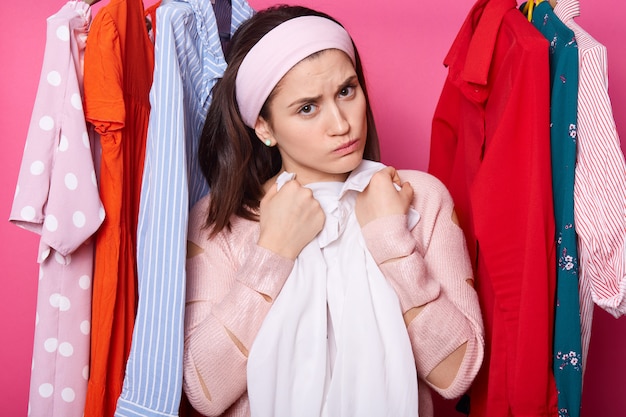 Discontent young female embraces white blouse. Beautiful woman wears rose sweater and hair band. Upset girl with lots clothes in wardrobe. Charming lady isolated over pink wall Fashion concept.