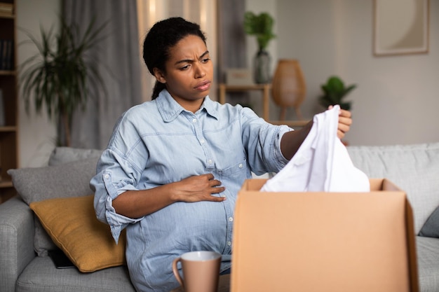 Discontented Pregnant Black Woman Holding Delivered Clothes Unpacking Box Indoor