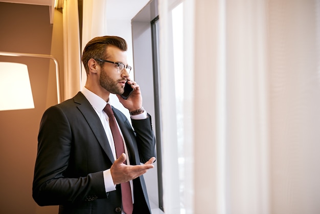 Discussing business topics at business trip. Close-up of spectacled businessman with smartphone standing near the window at hotel room. Close-up