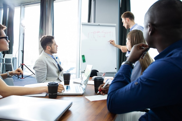 Discussing company progress. Confident young man standing near whiteboard and pointing graph while his colleagues sitting at the desk.