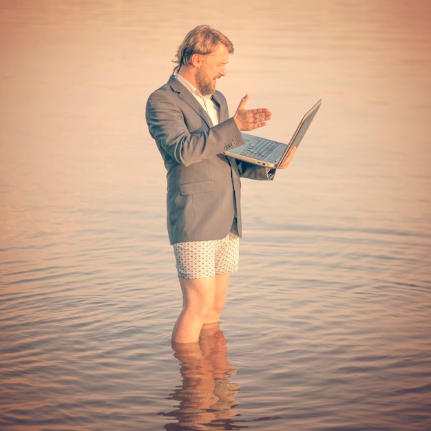 Disgruntled bearded man in a gray jacket and underwear with a laptop in hands standing kneedeep in water Toned