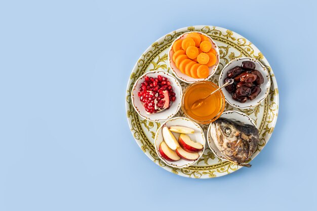 A dish with a New Year's treat for the New Year's holiday Rosh Hashanah pomegranate honey dates apple slices and carrots Blue background