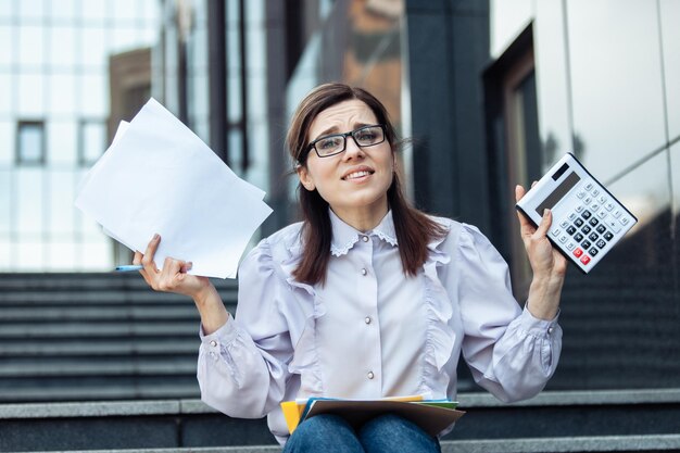 Disheartened business woman holding a calculator and paper sheets while sitting on the stairs Economist Lifestyle