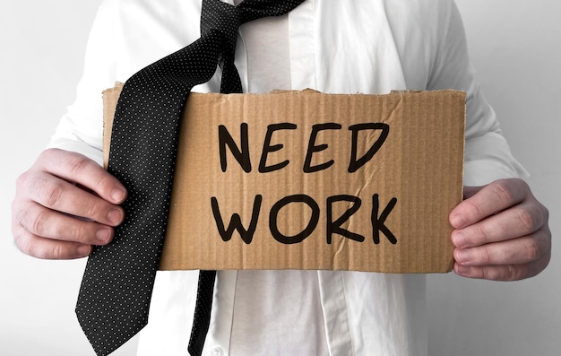 A disheveled business man holds up a cardboard sign with the words Need Work