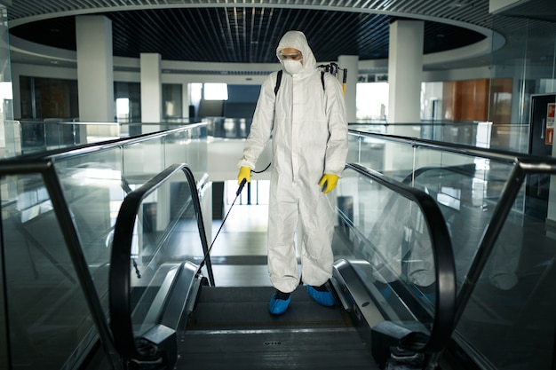 Disinfection worker wearing a protective suit professionally cleans up an escalator in an empty business center. A man equiped with antibacterial clothes sterilizes shopping mall. Covid concept.