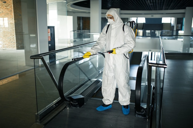 Disinfection worker wearing a protective suit professionally cleans up an escalator in an empty business center. A man equiped with antibacterial clothes sterilizes shopping mall. Covid concept.