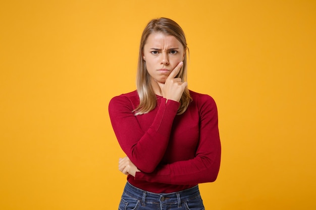 Displeased pensive young blonde woman girl in casual clothes posing isolated on yellow orange wall background studio portrait. People lifestyle concept. Mock up copy space. Put hand prop up on chin.