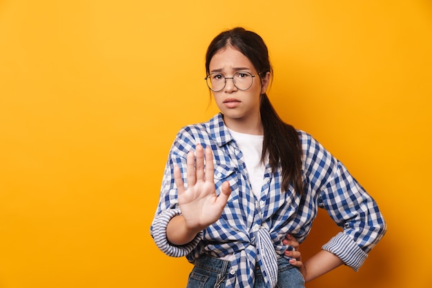 displeased sad cute teenage girl in glasses posing isolated over yellow wall  showing stop gesture.