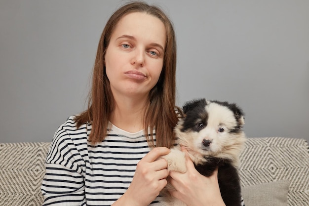 Displeased sad tired Caucasian woman wearing striped shirt sitting on couch with black and white pup