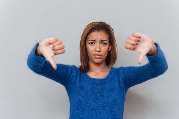 Displeased young african woman in sweater showing thumbs down.