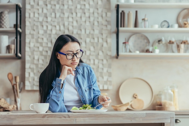 Displeased young asian woman in glasses and denim shirt sitting in the kitchen at home at the table