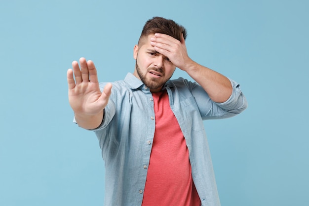 Displeased young bearded guy in casual shirt posing isolated on pastel blue background studio portrait. People lifestyle concept. Mock up copy space. Showing stop gesture with palm, put hand on head.