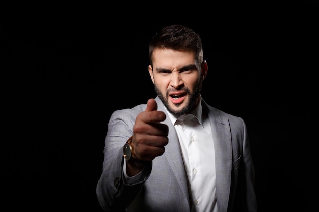 Displeased young successful businessman in suit ponting finger to camera on black