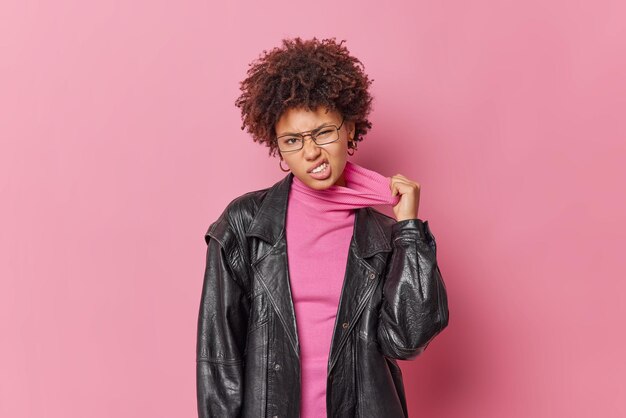 Displeased young woman has heatstroke stretches collar of turtleneck dressed in black leather jacket transparent glasses isolated over pink background. Annoyed curly haired female has cheeky look