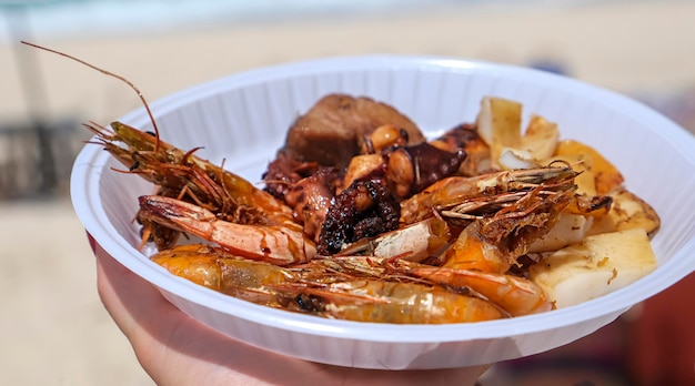 Disposable plate with a mix of grilled seafood shrimp, octopus, lobster, squid. BBQ exotic food on the beach. Selective focus. Low depth of field.