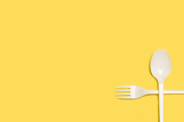 Disposable white plastic fork and spoon on a yellow background with copy space