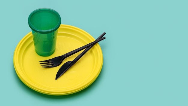 Disposable yellow plates, green glass for drinks, cutlery, on a green background. Still life with pl