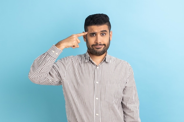 Dissatisfied angry businessman with beard showing crazy or stupid gesture holding finger near temple depressed has no ideas wearing striped shirt Indoor studio shot isolated on blue background