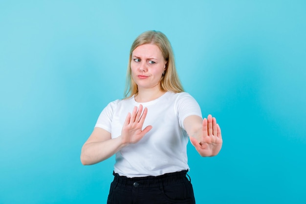 Dissatisfied blonde girl is showing stop gesture with hands on blue background