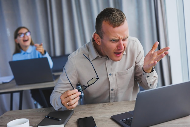 Dissatisfied businessman looking at something on smartphone while working The concept of a modern successful person Young confused guy in glasses sitting at desk in open space office with laptops