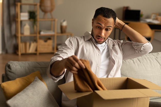 Dissatisfied buyer Frustrated black man unboxing package after delivery holding clothes sitting on couch at home