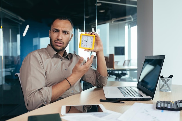 Dissatisfied hispanic man looking at camera and showing clock businessman frustrated waiting for