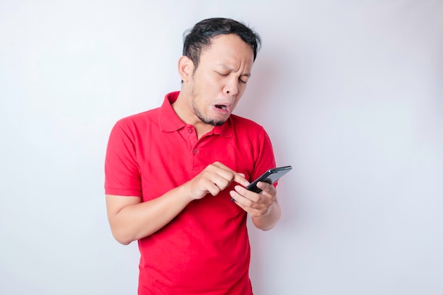 A dissatisfied young Asian man looks disgruntled wearing red tshirt irritated face expressions holding his phone