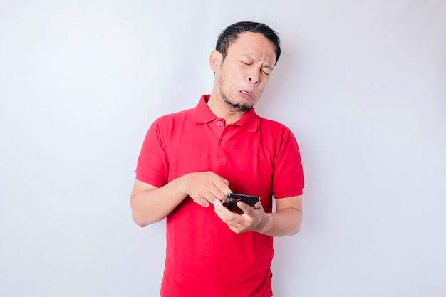 A dissatisfied young Asian man looks disgruntled wearing red tshirt irritated face expressions holding his phone