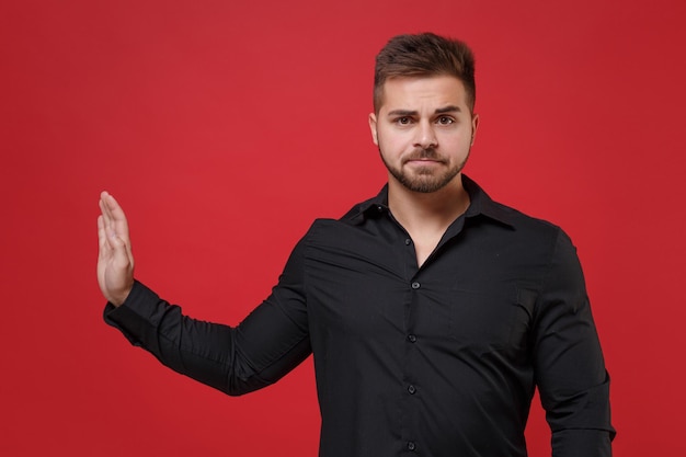 Dissatisfied young bearded guy 20s in classic black shirt posing isolated on red wall background studio portrait. People lifestyle concept. Mock up copy space. Showing stop gesture with palm aside.