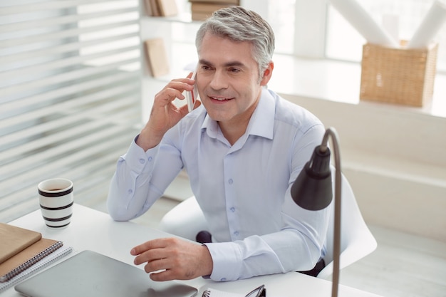 Photo distance communication. positive delighted nice man sitting at the table and making at call while working in the office