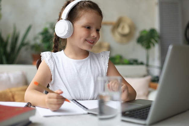 Distance learning. Cheerful little girl girl in headphones using laptop studying through online e-learning system.