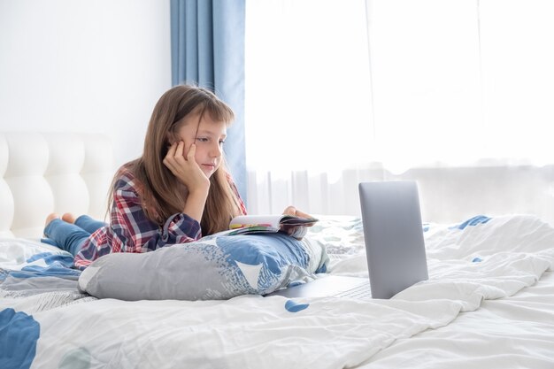 Distance learning online education. Schoolgirl studying with digital laptop notebook and doing school homework lying on bed at home