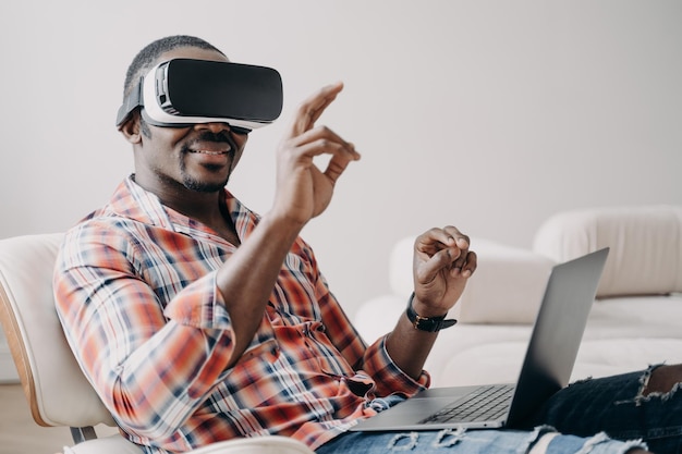 Distant work and futuristic gaming African american man in vr goggles working with laptop at home