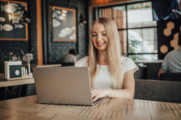 Distant work. A young blonde woman sits at a table in a cafe with a laptop and looks at the screen. Job search, online work