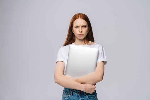 Distressed upset young business woman or student holding laptop computer and looking at camera on isolated gray background Pretty lady model with red hair emotionally showing facial expressions