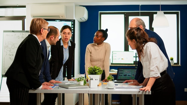 Diverse businesspeople brainstorming while meeting at company office standing at conference desk looking in documents. Colleagues working planning success financial strategy discussing in office