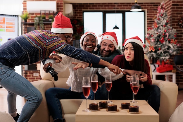 Diverse coworkers playing video games at christmas event, celebrating winter holiday festivity with colleagues at workplace decorated with seasonal ornaments. Having fun gaming at party.