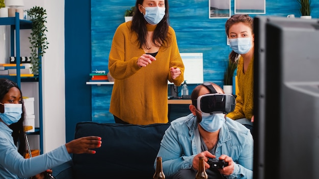 Diverse friends trying to win playing video games experiencing virtual reality headset having fun enjoying beer and popcorn wearing face mask to prevent spreading virus in time of global outbreak