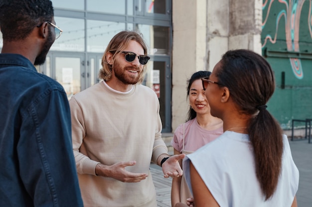 Diverse group of contemporary young people chatting outdoors in urban city setting, focus on smiling man wearing sunglasses