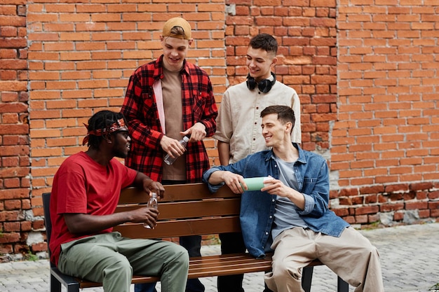 Diverse group of young guys wearing streetstyle fashion while sitting on bench and chatting against