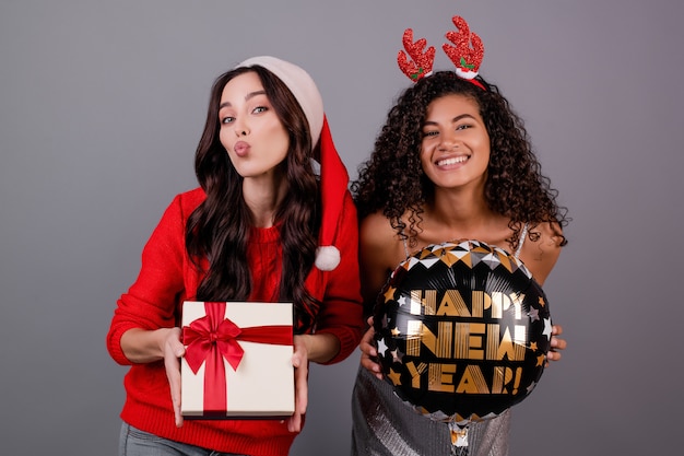 Diverse happy women with gift box and happy new year balloon wearing santa hat isolated over grey