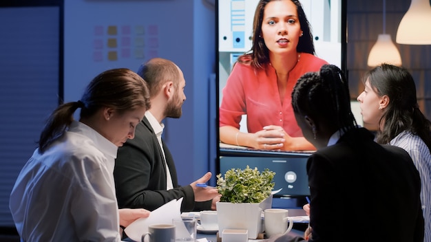 Diverse multi-ethnic businesspeople discussing with remote manager during online videocall conference meeting late at night in company office room