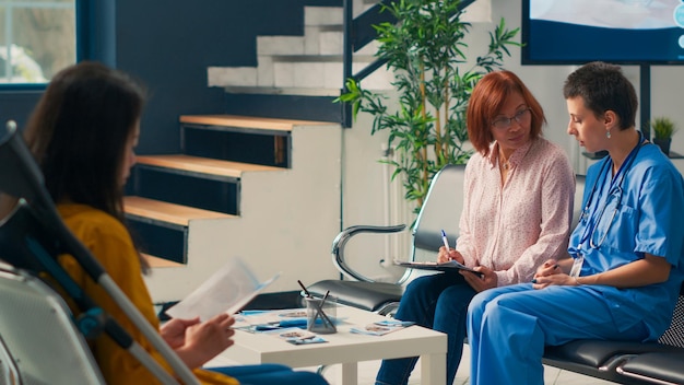 Diverse people filling in checkup report files in facility lobby, having medical appointment with physician at clinic. Nurse consulting elderly patient with insurance, healthcare support.