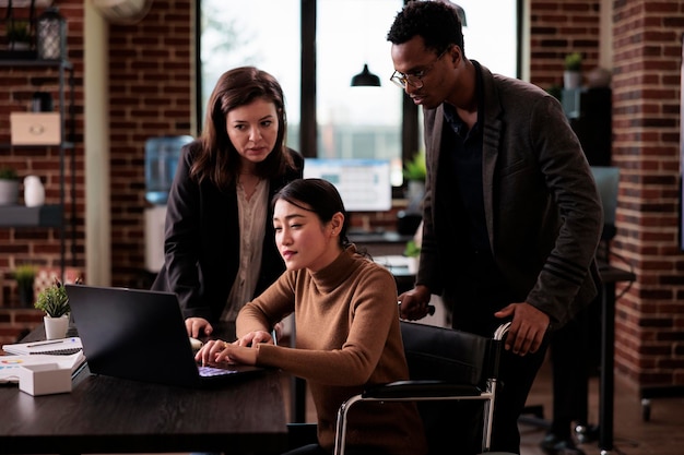 Diverse team of colleagues working with paralyzed employee sitting in wheelchair, suffering from chronic disability and impairment. Business people using laptop, health condition.