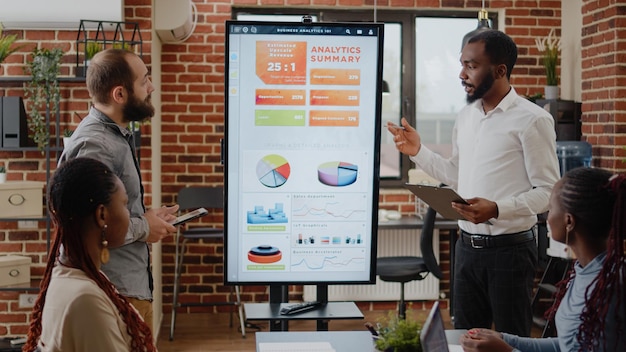 Diverse team of men explaining data analysis with charts on monitor. People talking about business growth and development, meeting with coworkers to do project presentation in startup office.